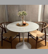 A white Carrara marble dining table with a bevelled edge on a slender pedestal base, accompanied by mid-century style wooden chairs.