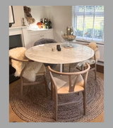 A white Carrara marble dining table with a bevelled edge on a slender pedestal base, accompanied by modern white chairs with wooden legs. The table is set in a bright room with large windows and a light interior.