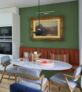 An oval Italian marble tulip dining table with a bevelled and tapered edge, placed on an aluminium lacquered stem base. The table is set in a room with green walls, a framed picture on the wall, and modern furniture around it.