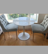 A white Carrara marble dining table with a bevelled edge on a slender pedestal base, accompanied by modern grey chairs with wooden legs. The table is set in a bright room with large windows and a light interior.