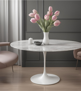 Round marble dining table with a central aluminum base, presented in a choice of sizes. The table is set in a room with two beige chairs, and there is a vase with tulips on the table.