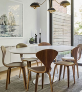 A white tulip dining table with a wooden top and an aluminum base, surrounded by wooden cherner style chairs, in a room with a large window and a painting on the wall.